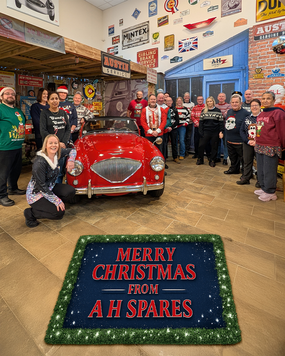 A H Spares staff in Christmas jumpers with a red Austin Healey and Merry Christmas message inside classic car workshop