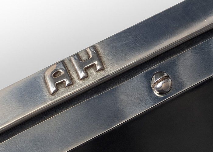 Close-up of a polished metal Austin Healey aero screen engraved with 'AH' initials, featuring a clean, fingerprint-free surface and a slotted chrome screw.